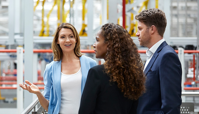 Une femme d’affaires d’âge mûr discute avec des collègues dans une usine de fabrication d’automobiles. Une entrepreneure affiche un sourire alors qu’elle se tient aux côtés de collègues près d’une rampe. Ces personnes portent une tenue de ville dans l’usine.